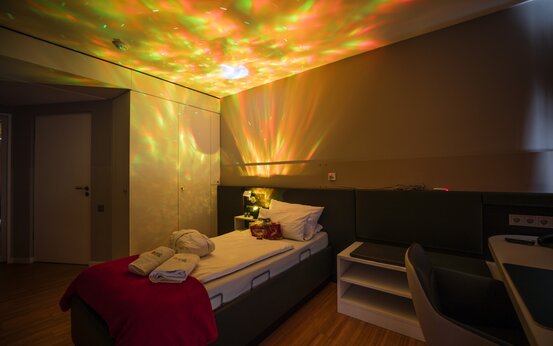 Modernes Einzelzimmer mit beleuchtetem Pflegebett, bunten Deckenlichtern, Handtüchern und Bademantel