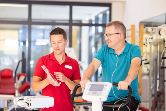 Physiotherapeut erklärt Patient auf Ergometer mit Elektroden das Training im hellen Rehazentrum