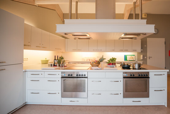 Moderne, geräumige Lehrküche mit zwei Elektroherden, weißen Schränken, Kochfeld und frischen Zutaten auf den Arbeitsflächen