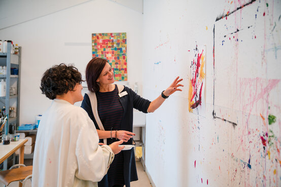 Therapeutin bespricht mit Patientin ein abstraktes Kunstwerk im hellen, kreativ gestalteten Therapieraum der Klinik