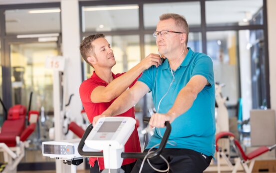 Älterer Mann mit Sauerstoffbrille trainiert auf Ergometer, betreut von Physiotherapeut