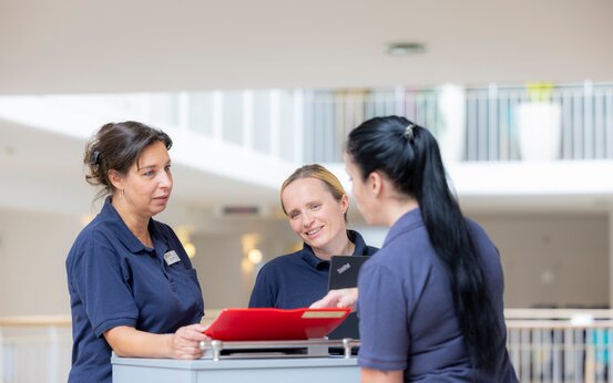 Drei Pflegekräfte in blauer Dienstkleidung besprechen Patientenunterlagen an einem mobilen Schreibtisch