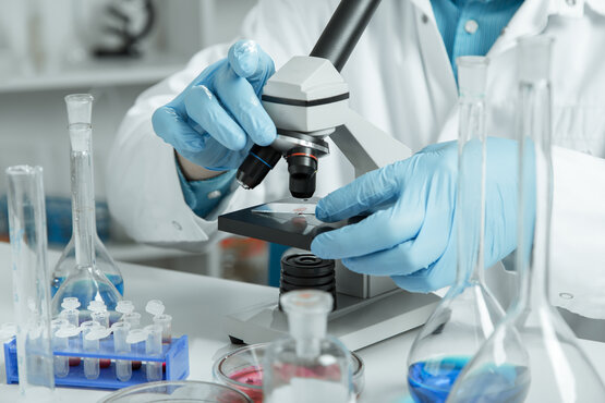 Nahaufnahme eines Wissenschaftlers in weißem Laborkittel und blauen Handschuhen beim Arbeiten am Mikroskop