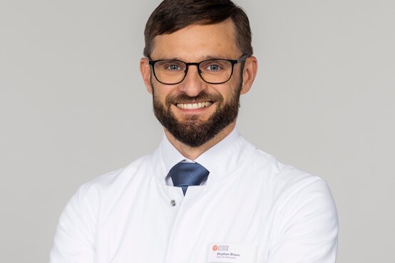 Stephan Braun. Porträt eines männlichen Arztes mit kurzem braunem Haar, Bart und Brille im weißen Kittel und blauer Krawatte