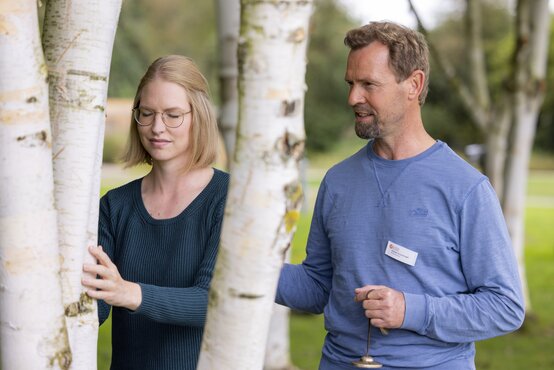 Blonde Frau mit Brille berührt mit geschlossenen Augen einen Baumstamm, angeleitet von Therapeut im Park