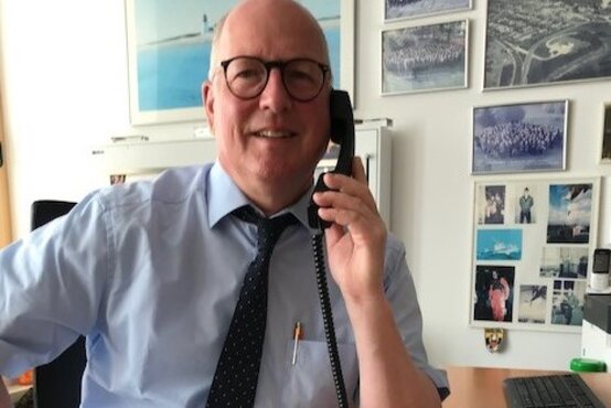 Älterer, lächelnder Mann mit Glatze, Brille, blauem Hemd und Krawatte telefoniert am Schreibtisch