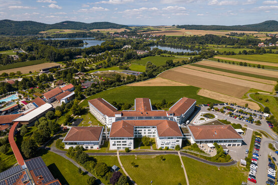 Luftaufnahme der Schön Klinik Bad Staffelstein mit roten Dächern, umgeben von Grünflächen, Parkplätzen, Feldern und einem See