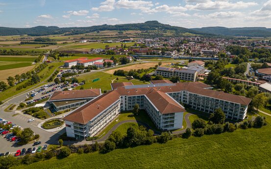Luftaufnahme der Schön Klinik Bad Staffelstein mit rotem Ziegeldach, umgeben von Wiesen, Bäumen und Parkplätzen
