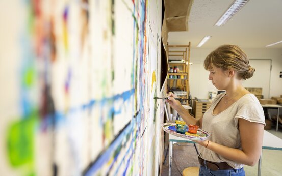 Junge Frau malt mit Pinsel an bunter Leinwand im hellen Kunstatelier, Kunsttherapie und kreative Entfaltung