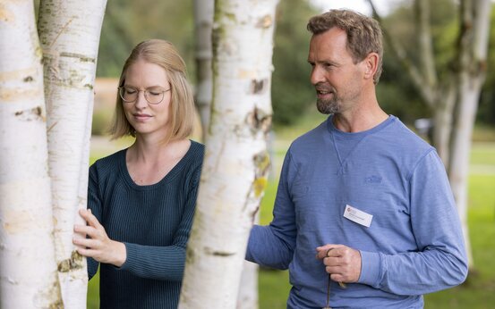 Junge Frau mit Brille berührt mit geschlossenen Augen Birkenstamm, älterer Mann mit Glocke im Park bei Achtsamkeitsübung