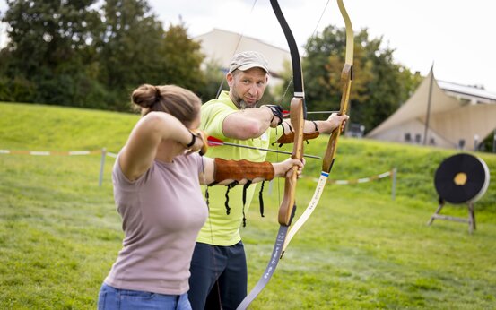 Frau erhält Anleitung beim therapeutischen Bogenschießen im Freien, Zielscheibe und Trainer im Hintergrund