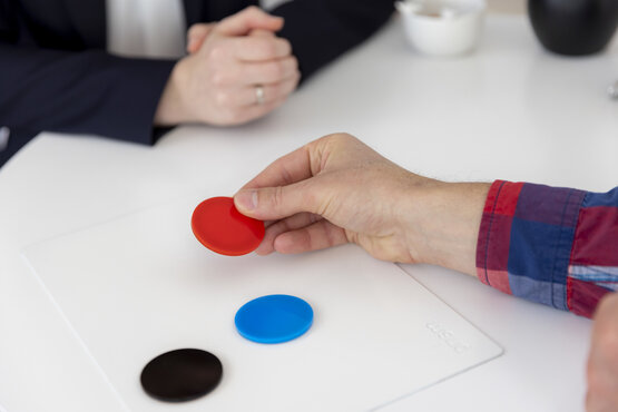 Psychotherapeutische Sitzung mit zwei Personen, Hände am Tisch, rote, blaue und schwarze Markerscheiben als Hilfsmittel