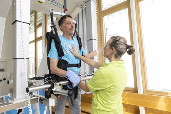 Älterer Mann bei der Gangrehabilitation mit robotischem Therapiegerät, unterstützt von Physiotherapeutin