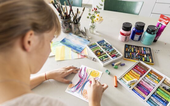 Frau zeichnet mit bunten Ölkreiden an einem Tisch mit Pastellkreiden, Pinseln, Acrylfarben und Blumen
