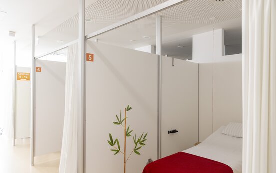 Helles Behandlungszimmer mit Einzelbett, roter Decke, weißen Trennwänden, Kabinennummern und Zimmerpflanze