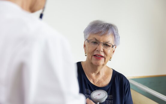 Ältere Frau mit Brille sitzt im Behandlungszimmer, Arzt misst Blutdruck