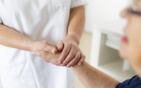 Pflegerin in weißer Kleidung hält fürsorglich die Hand einer älteren Frau im Pflegebett
