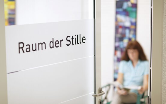 Glas-Tür mit Aufschrift „Raum der Stille“, Blick in hellen Ruheraum mit Frau beim Lesen und farbigem Wandbild