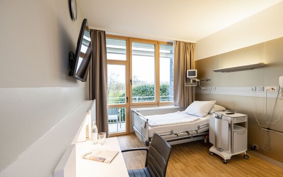 Modernes Komfortzimmer in Klinik mit elektrisch verstellbarem Bett, Holzfußboden, bodentiefen Fenstern und Schreibtisch