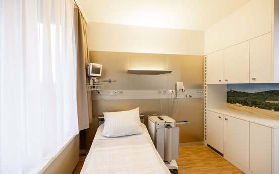 Modernes Komfortzimmer in Klinik mit großem Fenster, Einzelbett, Nachttisch, Holzboden