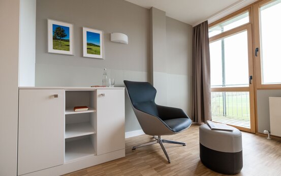 Modernes Komfortzimmer in Klinik mit Holzboden, großer Fensterfront, Sessel, Hocker und Sideboard
