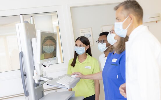 Ärzteteam und Pflegekräfte mit Masken besprechen Patientendaten am Computer im hellen Klinikflur