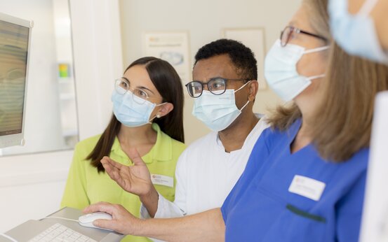 Drei medizinische Fachkräfte mit Mundschutz betrachten gemeinsam Computerbildschirm in heller Klinik