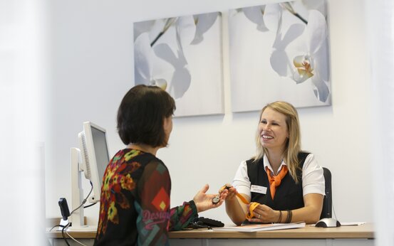 Mitarbeiterin mit Namensschild übergibt Patientin im modernen Servicebüro einen Schlüssel