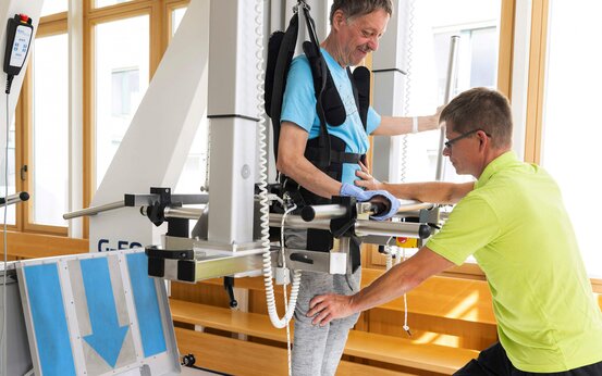 Älterer Mann trainiert mit Therapiegurt an Laufroboter, unterstützt von Physiotherapeut