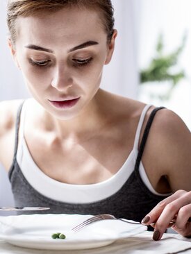 Junge Frau blickt ernst auf fast leeren Teller mit wenigen Erbsen, Messer, Gabel und Glas Wasser auf hellem Esstisch
