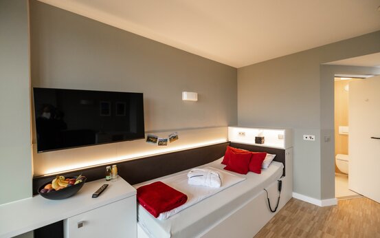 Modernes Einzelzimmer in Klinik mit Einzelbett, Bademantel, rotem Handtuch, Flachbildfernseher und offenem Bad