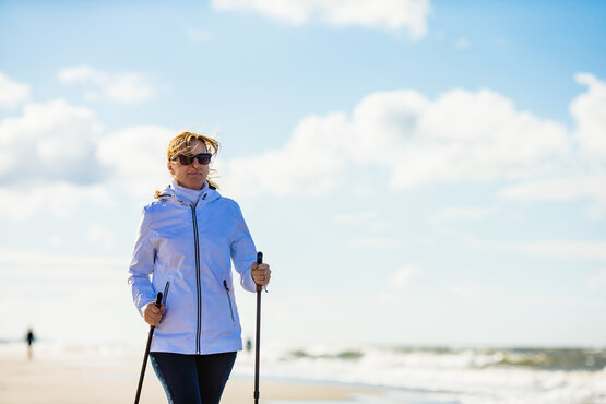 Frau in Sportkleidung geht mit Nordic-Walking-Stöcken am Strand, Meer und Himmel im Hintergrund