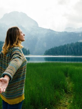 Frau mit ausgestreckten Armen am See, umgeben von grüner Natur
