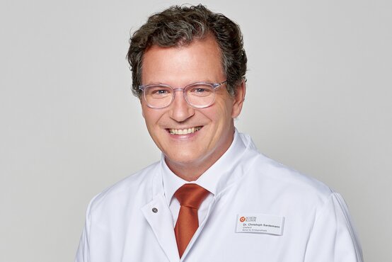 Porträt eines lächelnden Arztes mit lockigem braunem Haar, Brille und weißem Kittel