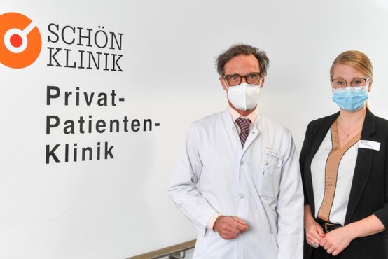 Zwei Mitarbeitende der Schön Klinik Düsseldorf, Arzt im weißen Kittel und Frau im Business-Outfit, mit Masken vor Klinik-Logo