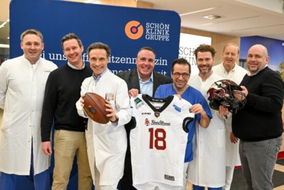 Acht Männer, darunter Ärzte in weißen Kitteln, halten Football, Trikot und Helm vor Schön Klinik Logo – Kooperation mit Rhein Fire