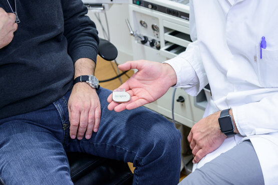Ein Arzt im weißen Kittel zeigt einem Patienten einen Zungenschrittmacher im Untersuchungszimmer.