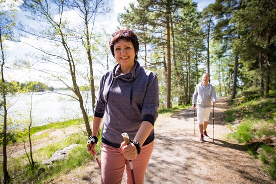 Zwei Erwachsene beim Nordic Walking auf sonnigem Waldweg am See, Frau lächelt, grüne Natur