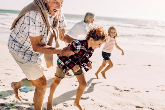 Älterer Mann läuft lachend barfuß mit Enkel am Strand, Familie bewegt sich aktiv bei Sonnenschein