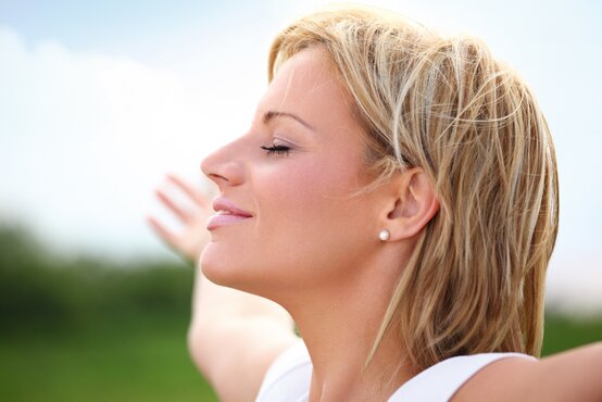 Lächelnde Frau mit geschlossenen Augen atmet tief ein, Arme ausgestreckt, im Freien vor grünem Hintergrund