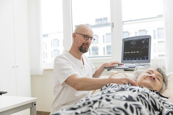 Arzt führt eine Ultraschalluntersuchung am Hals einer älteren Patientin durch, Ultraschallgerät im Hintergrund