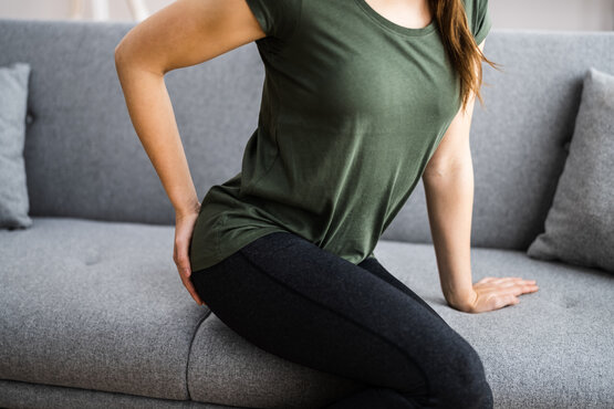 Junge Frau sitzt auf grauem Sofa und hält sich den Gesäßbereich