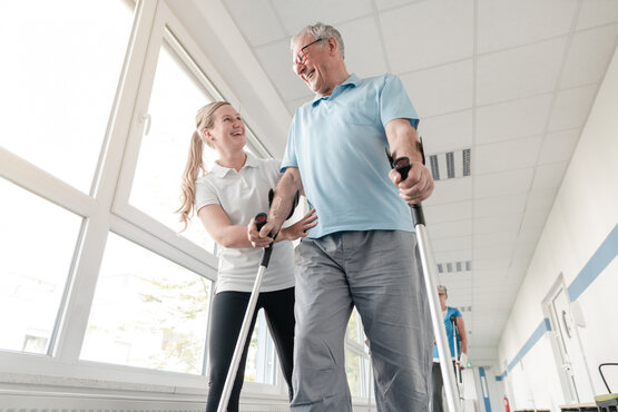 Älterer Mann mit Gehhilfen wird von Physiotherapeutin beim Gehtraining im hellen Klinikflur unterstützt