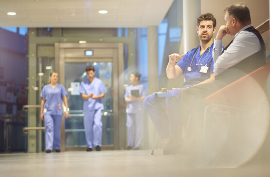 Mehrere Pflegekräfte in blauer Krankenhauskleidung gehen durch modernen Flur; Arzt spricht mit Mann auf Bank