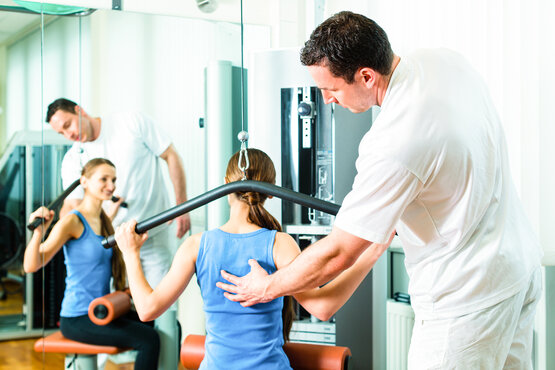 Physiotherapeut unterstützt junge Frau beim Rückentraining an Fitnessgerät