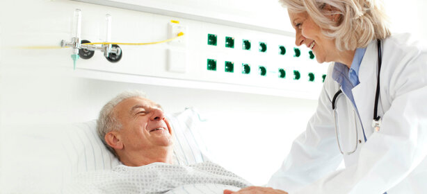 Ärztin im weißen Kittel lächelt älterem Patienten im Krankenhausbett zu, moderne Klinikzimmer-Ausstattung im Hintergrund