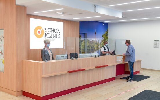 Moderner, barrierefreier Empfangsbereich der Schön Klinik Düsseldorf mit Holztheke, Plexiglas, Masken und Computerarbeitsplätzen