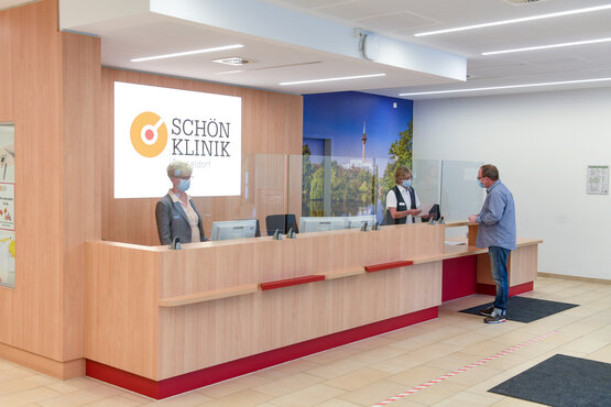 Moderner, barrierefreier Empfangsbereich der Schön Klinik Düsseldorf mit Holztheke, Plexiglas, Masken und Computerarbeitsplätzen