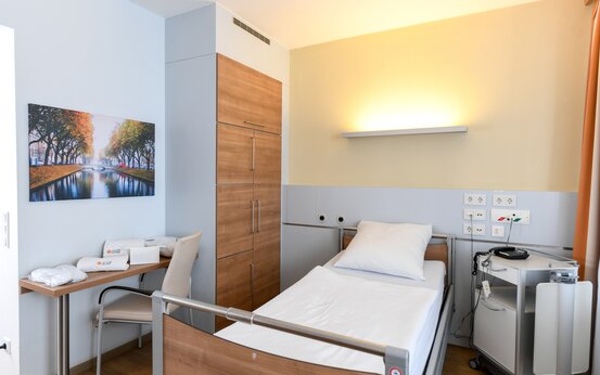 Klinik-Einzelzimmer mit höhenverstellbarem Bett, Schreibtisch, Nachttisch und warmem Licht