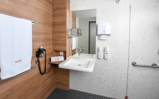 Barrierefreies Klinik-Badezimmer mit unterfahrbarem Waschbecken, Haltegriffen, großem Spiegel und modernem Holzdekor